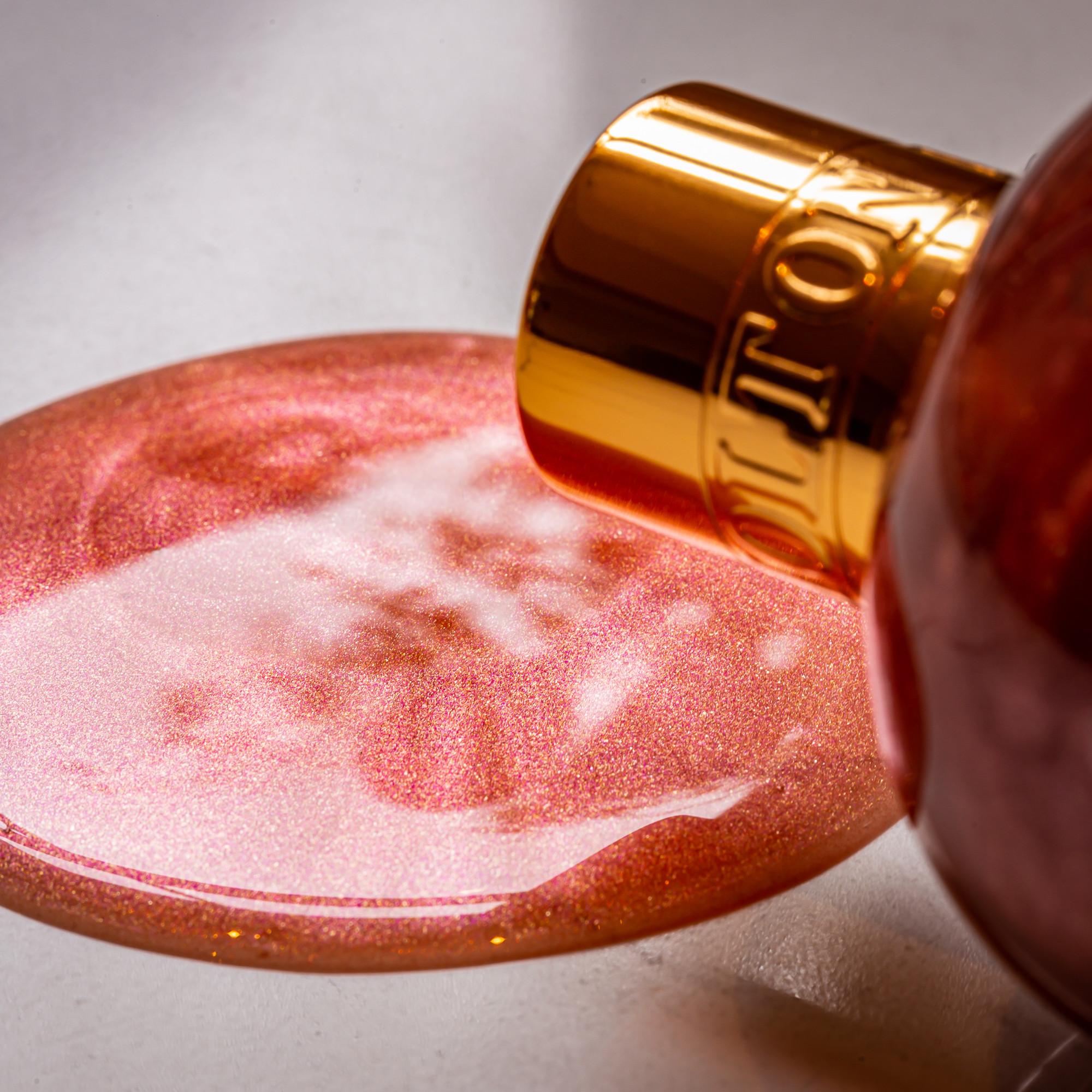 A close-up of the Rose Dunes Bath & Shower Gel 300ml with its gold cap off, pouring shimmery pink rose-infused gel onto a smooth surface, as sparkles and light glimmer in the liquid.