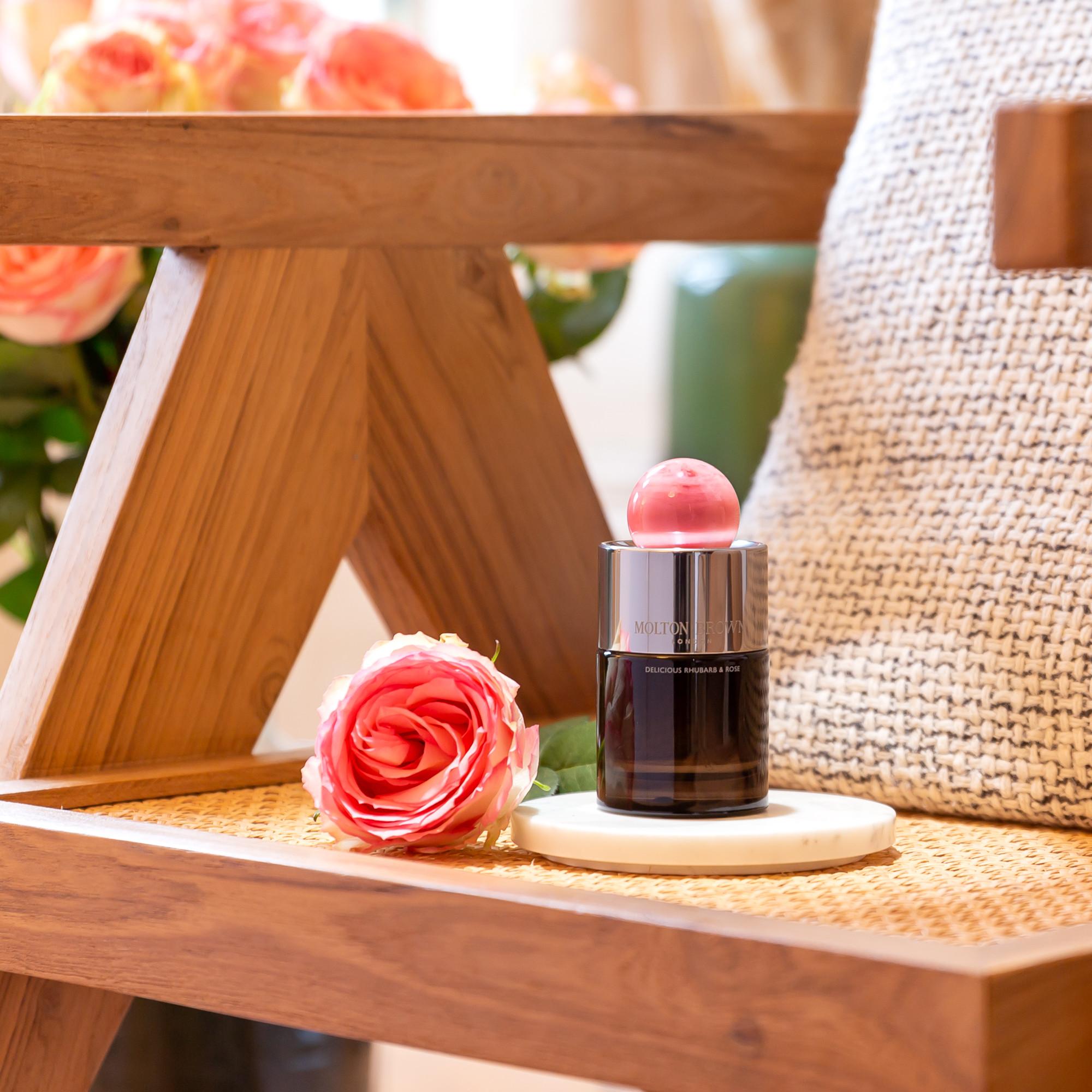 A pink rose and a bottle of Delicious Rhubarb & Rose Eau de Parfum 100ml with a pink cap rest on a wooden chair. A pillow and a bouquet of pink roses in the background add to the cozy, elegant perfume setting.