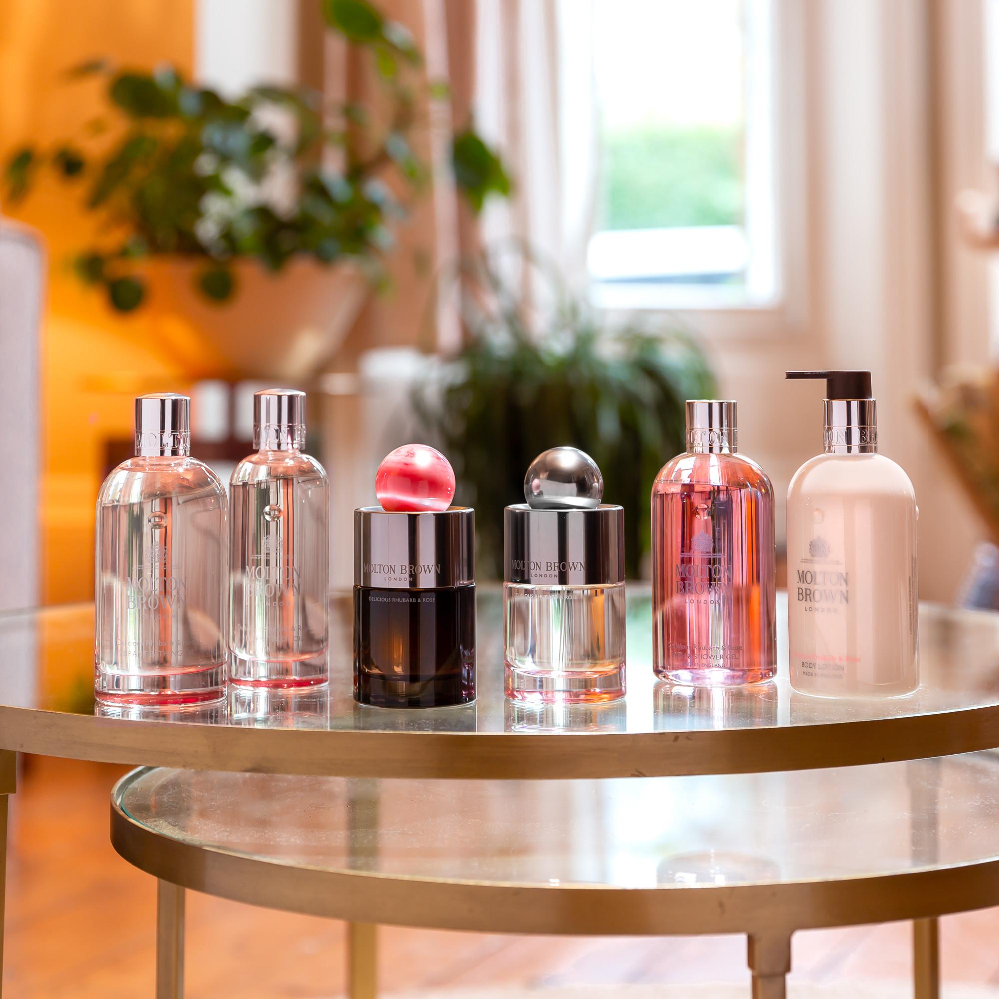 Five bottles of Molton Brown, including the Delicious Rhubarb & Rose Eau de Parfum 100ml and a lotion, are displayed on a glass table with plants and sunlight visible in the blurred background.