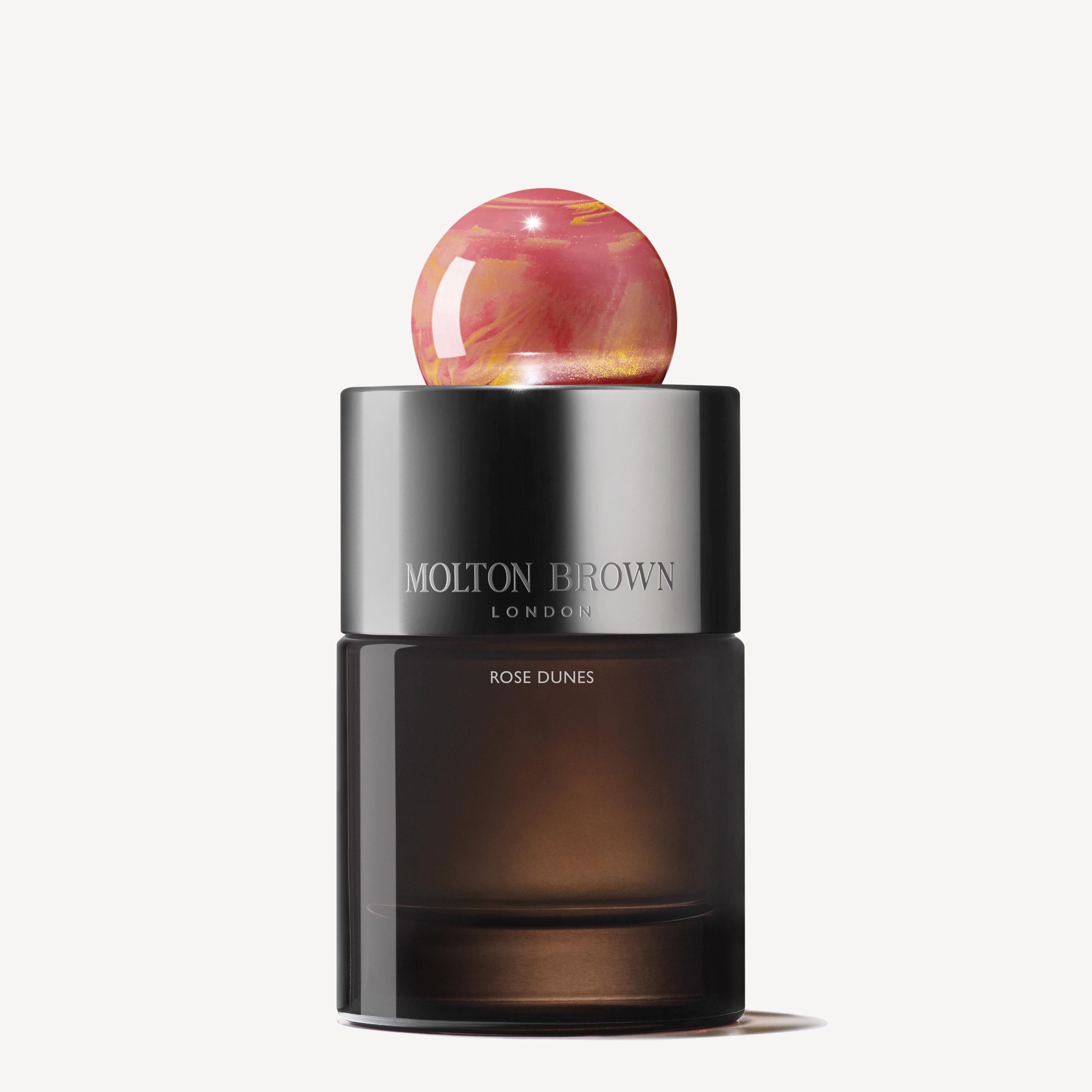 A dark glass bottle of Rose Dunes Eau de Parfum 100ml, with a metallic cap and a round pink marbled stopper, evokes Arabian desert mystique against a plain white background.