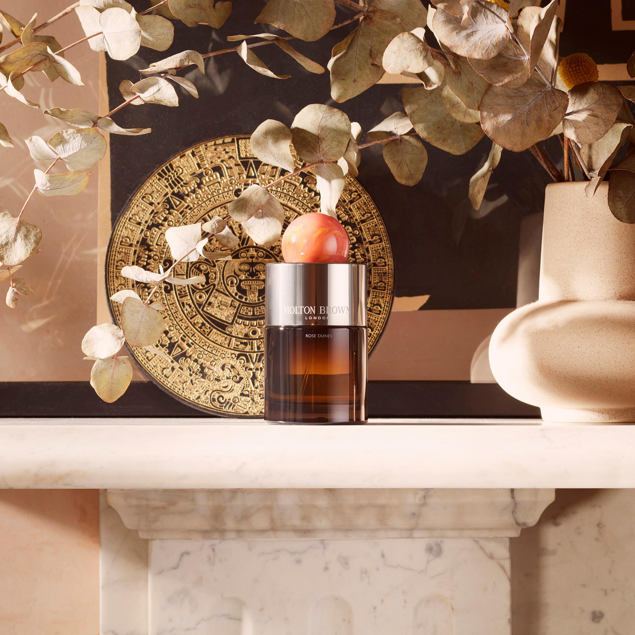 A 100ml Rose Dunes Eau de Parfum bottle, made of brown glass with a round peach cap, rests on a white marble mantel amid dried eucalyptus, a decorative gold disc, and a beige vase.