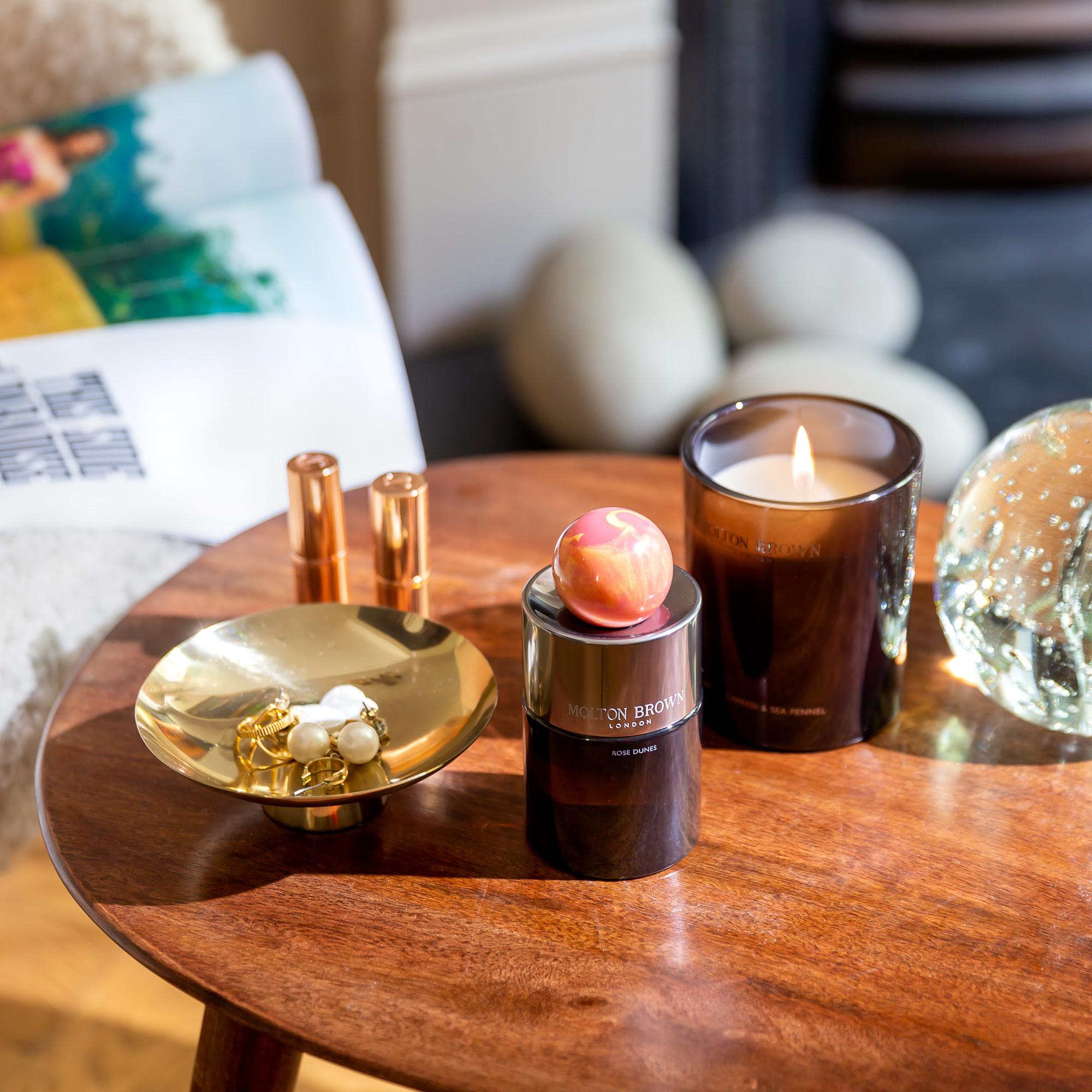 A wooden table holds a lit candle, a jar with a decorative ball, a gold jewelry dish, two lipsticks, Rose Dunes Eau de Parfum 100ml, and a glass orb. An open magazine is on a chair nearby.