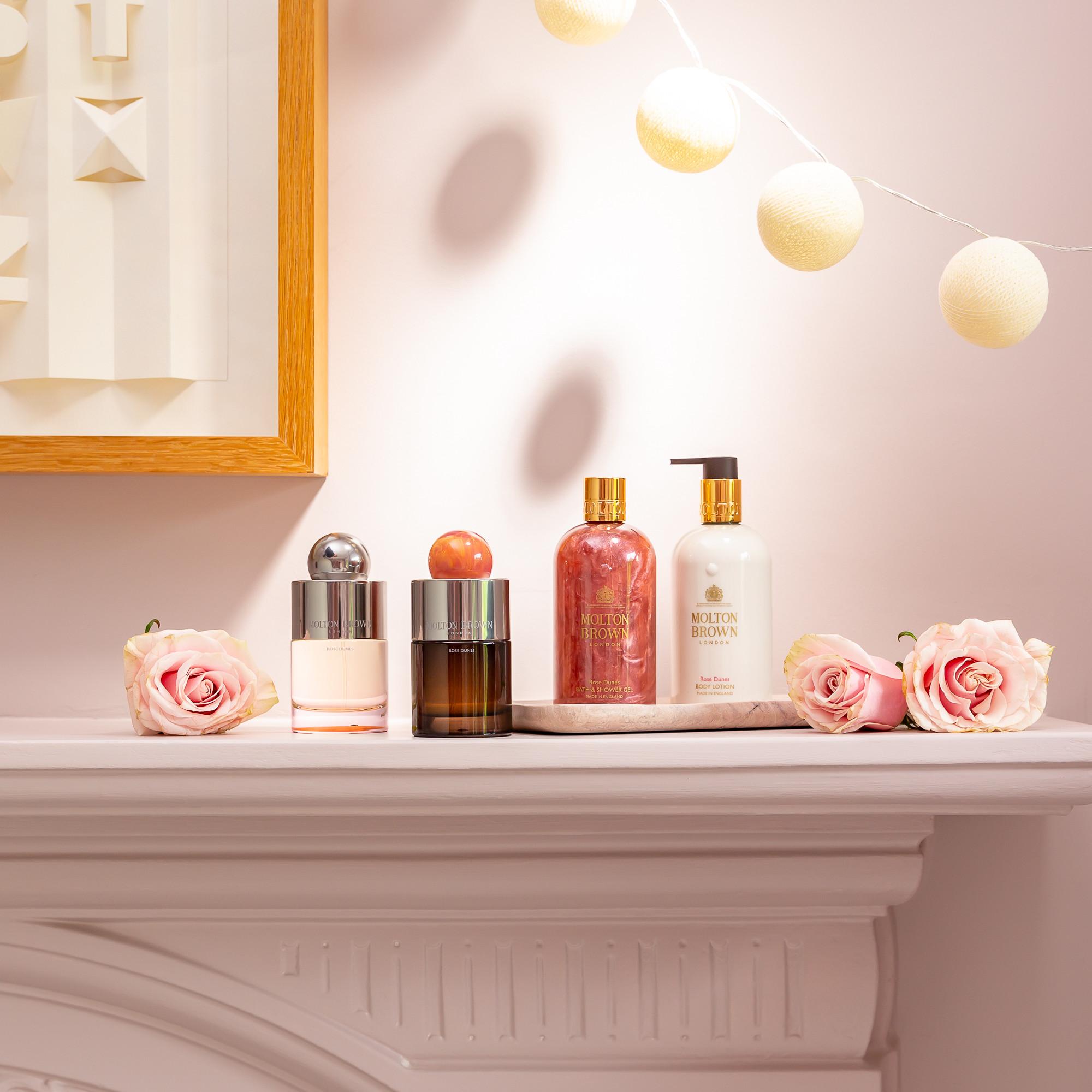 A white mantel decorated with pink roses, string lights, and luxury cosmetics—including Molton Brown hand soap, lotion, and Rose Dunes Eau de Parfum 100ml—stands against a soft pink wall adorned with framed art.