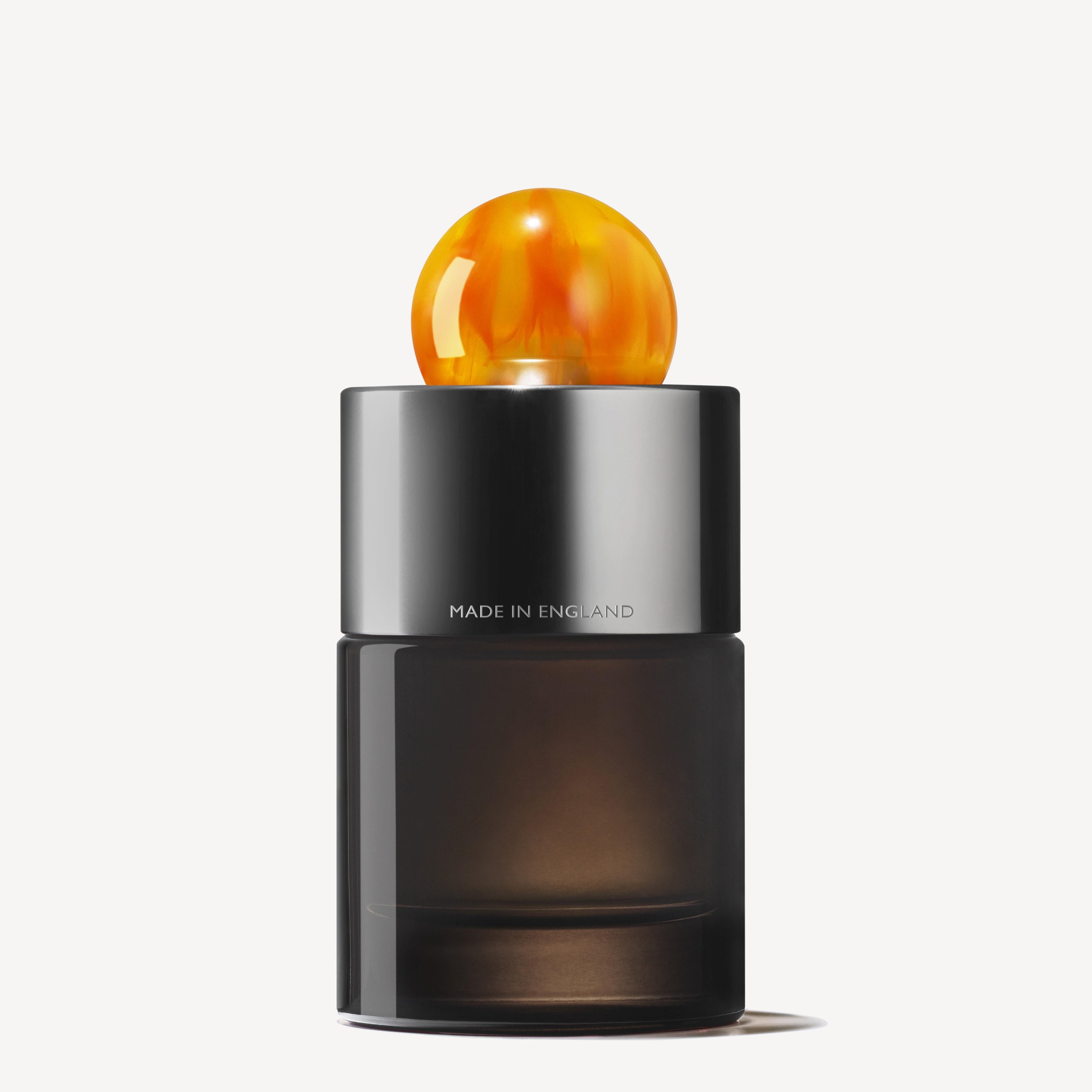 The Sunlit Clementine & Vetiver Eau de Parfum 100ml features a sleek, dark glass bottle with a shiny silver collar printed with "MADE IN ENGLAND" and a round amber cap, embodying elegance against a plain white background.