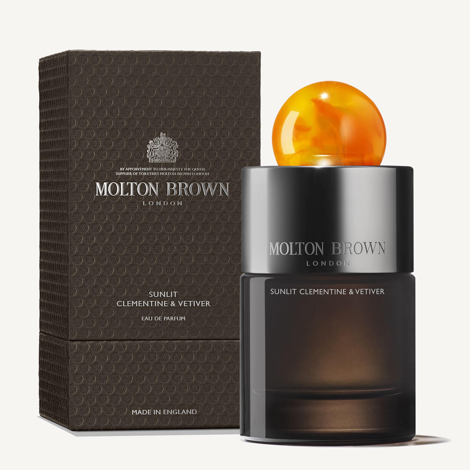 A 100ml bottle of Sunlit Clementine & Vetiver Eau de Parfum with a round orange cap stands beside its textured brown box with silver lettering.