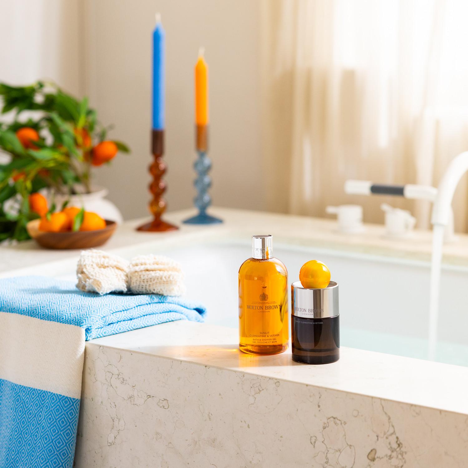 Two bottles of luxury bath products, including Sunlit Clementine & Vetiver Eau de Parfum 100ml, sit on the marble tub’s edge with a lemon resting on one. Nearby, blue towels, crochet mitts, fresh oranges, and two lit candles evoke a spa-like ambiance.